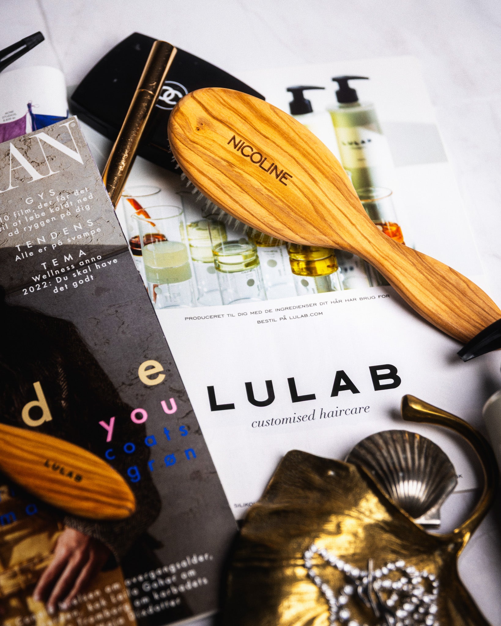 Personlig hårbørste med indgraveringen ‘NICOLINE’ placeret oven på brochurer, omgivet af makeup, hårprodukter og dekorative genstande.
